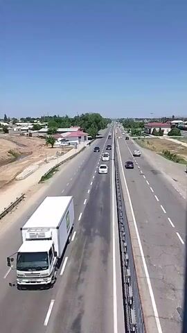 The video shows a view from an overpass looking down onto a highway with multiple lanes in both directions. Cars are driving on the highway. On the left side of the highway, there's a white truck with "HAVAS" printed on its side. Beyond the highway, there's a landscape with buildings and trees. The sky is clear and blue.