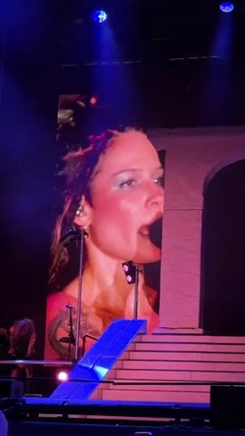 A video of a concert performance features a large screen displaying a woman in a corset and underwear. The woman on screen sings and dances while a spotlight shines on her. On the stage below, another woman plays a guitar. The stage is decorated with pink arches and stairs, and the lighting is predominantly pink and purple.