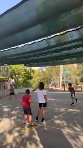 The video shows a basketball court with several children.  One child wears a red shirt and dark shorts while standing inside of a yellow hoop.  Another child wears a white shirt and dark shorts while standing next to the child in the hoop.  Other children are also on the court; one near the basketball hoop, and another near the edge of the court.  The court is surrounded by trees and other greenery.  The court is covered by a green mesh canopy.  The video is shot from under the canopy.  The audio includes children talking and making noise.