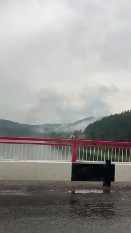 A view from a bridge over a body of water with mist rising from forested hills in the background. The bridge has a white barrier with red railings and a black object is attached to the railing. The road surface in the foreground is wet with puddles. The sky is cloudy and gray.