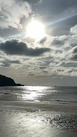 The video shows a beach scene. The sun is bright and partially obscured by clouds, casting a glare on the water. There are some dark clouds in the sky. In the distance, a dark, hilly coastline is visible on the left. The ocean has gentle waves, and the wet sand reflects the sunlight. Some small puddles of water are present on the sand in the foreground. A small dark boat is visible in the distance on the water. There are no people clearly visible or identifiable in the footage. The overall impression is a dramatic, possibly overcast, beach landscape.