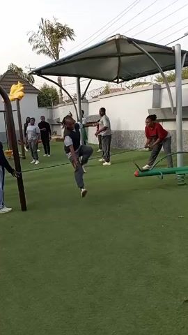A group of people are gathered in an outdoor playground area with green artificial turf. There is a covered structure supported by metal poles, and playground equipment like a seesaw and a bar structure with yellow decorative tops. Some individuals are standing and watching, while others are actively participating in some kind of rope course activity. People are walking and jogging around the playground. Two people are seen attempting to jump or step over a rope strung between poles, displaying enthusiastic body language. One of the participants is wearing red pants and white sneakers, while the other is in dark athletic pants and white sneakers. The atmosphere appears to be light and fun.