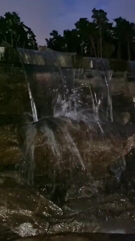 The video captures a nighttime view of a man-made waterfall cascading over rocks. The water appears illuminated, reflecting the surrounding lights, possibly from artificial sources within the landscape. The backdrop features a dark silhouette of pine trees against a twilight sky with visible clouds. The overall atmosphere is serene and natural, with the sound of flowing water present.