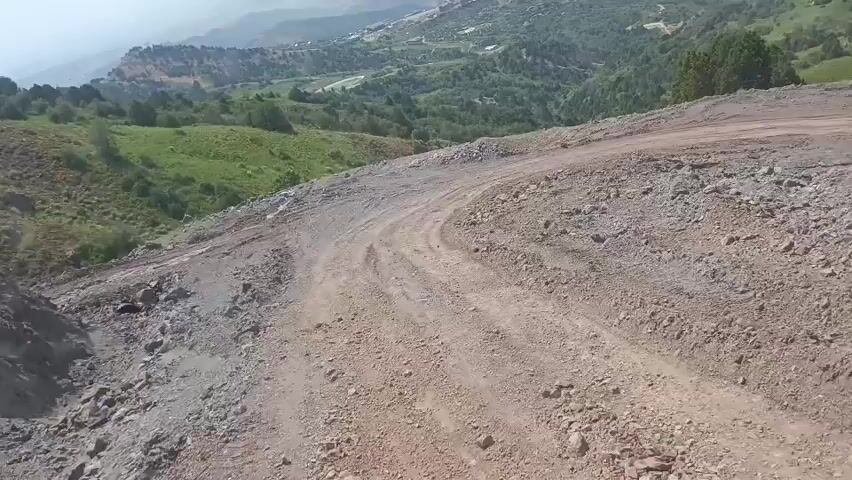 The video shows a view from a dirt road on a mountainside. The road is rough and rocky, with tire tracks visible. The surrounding landscape is mountainous and covered with green vegetation, including trees and grass. In the distance, there are buildings and roads, suggesting a settlement or resort area. The sky is hazy.
