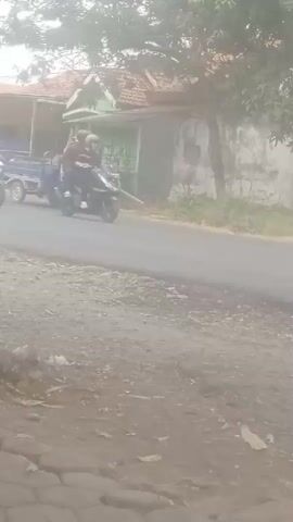 The video captures a street scene with several motorcycles and a three-wheeled vehicle. In the foreground, a dusty road and some pavement are visible. In the background, a modest house with a tiled roof and trees line the street.

At the beginning of the video, a blue three-wheeled vehicle with a cargo bed is seen moving along the road, driven by a person wearing a light-colored hat and casual clothing. Another motorcycle is visible to the right of the three-wheeled vehicle.

As the video progresses, the three-wheeled vehicle continues to move forward. A second motorcycle, ridden by two people, passes by on the right. A third motorcycle, ridden by a single person wearing a mask and helmet, is also seen on the road. The scene appears to be in a rural or semi-rural area, with the light suggesting it is daytime. The focus is on the movement of these vehicles and their occupants along the road.