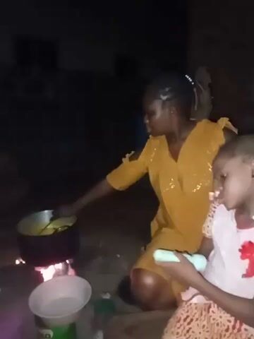 Here is a description of the video:

Two people, likely a mother and child, are outside near a cooking fire. The adult woman is wearing a yellow dress and stirring something in a large pot over a fire.  She has her hair pulled back and is looking at the pot.  The child is sitting next to her, wearing a white dress with a reddish pattern. The child is holding a small white object, possibly a bottle or toy, and is looking down. The environment is dark and the fire provides the main source of light. A building is visible in the background. The cooking setup appears to be a rudimentary fire on the ground. There are other containers near the fire.