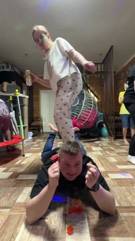 The video shows a group of people indoors, seemingly in a home environment with wooden floors and a staircase. A man is lying on his stomach on the floor with several small orange objects arranged in front of him. A woman in pajamas is standing on his back, holding a beverage can. A young boy in a yellow t-shirt is standing to the side, appearing to sing or speak into something resembling a microphone. Another child is partially visible on the staircase. The room also contains items such as a cat tree, a red trampoline, and a hanging chair.