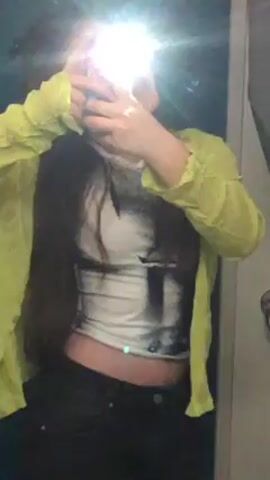 A person stands in front of a mirror, holding a phone up to take a picture of themself. The person is wearing a white and black patterned crop top, black pants, and a lime green long-sleeved sheer shirt. The camera flash reflects off the mirror, obscuring the person's face. The person's long dark hair hangs down their chest. The background is a white wall and a door frame is visible.