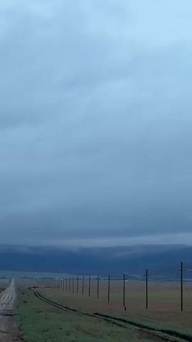 The video shows a long, straight dirt road stretching into the distance under a heavily overcast sky. The road appears to be muddy in places, and it is bordered on both sides by fields of green and brown vegetation. A line of wooden utility poles with wires runs parallel to the road on one side. In the distance, there is a dark, low-lying mountain range obscured by a layer of fog or low clouds. The overall scene conveys a sense of isolation and gloomy weather.