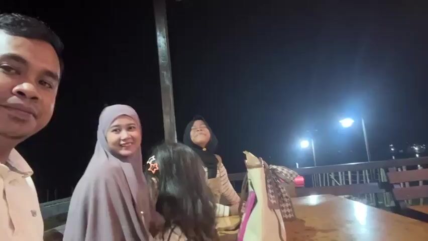 A group of four people are gathered at a table outdoors at night. The person on the far left is an adult male with short dark hair, wearing a light-colored collared shirt. To his right is an adult female wearing a light purple hijab and a light-colored top. In front of the woman is a young girl with dark hair, wearing a light-colored striped shirt and overalls, with a star-shaped clip in her hair. To the right of the young girl and behind her is another young female wearing a black hijab and a striped top. The group is seated at a wooden table with various items on it, including a bag. In the background, there is a railing, wooden chairs, and a dimly lit structure with lights. The overall lighting suggests it is nighttime, with a streetlight visible in the distance. The individuals appear to be posing for a selfie.