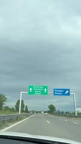 The video captures a first-person view from inside a vehicle driving on a multi-lane highway in Radinovo, Bulgaria, under an overcast sky.

The initial frame prominently displays an overhead green highway sign with two upward-pointing arrows. The sign reads "Свиленград" (Svilengrad) and "Бургас" (Burgas) in both Cyrillic and Latin script. To the right, a blue sign is partially visible, showing "Пловди" (likely Plovdiv) in Cyrillic. The car continues to drive forward, and the signs move out of view.

The highway features multiple lanes, with other cars and trucks visible in the distance. Guardrails line the sides of the road, and green trees and vegetation can be seen beyond them. A bridge or overpass is visible further down the road. The overall impression is of a routine journey on a cloudy day. Upbeat music is playing in the background throughout the video.
