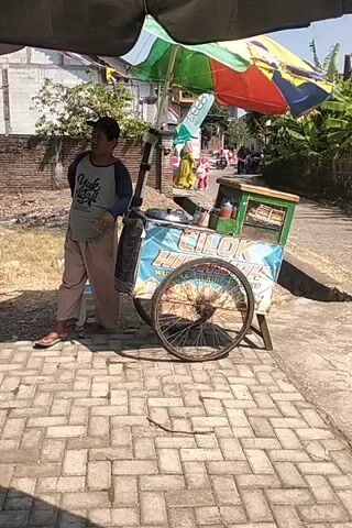 A man stands in front of a food cart. The cart has a blue tarp with writing on it and a large bicycle wheel. The man is wearing a grey long-sleeved shirt with writing on it and tan pants. He is looking towards the camera and smiling. Behind the food cart, there is a brick wall and some trees. The ground is paved with bricks. The video is taken from a low angle.