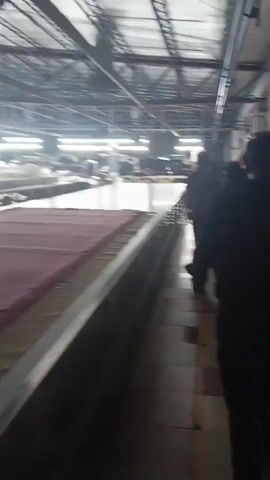 A group of men walk in a line down a hallway in what appears to be a factory. They are walking past a long table on their left, which has fabric laid out on it. The hallway is lit by overhead fluorescent lights. The men are all wearing dark clothing. The camera is following the men from behind.