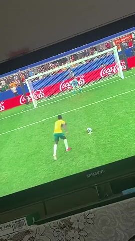The video shows a soccer game being played on a large screen television. In the foreground, a person is filming the television screen with a mobile phone. The television displays a soccer match in progress. One player, wearing a yellow jersey with the number 11, is in focus in the center of the screen. He appears to be reacting to a play. In the background of the televised game, another player in a light blue jersey is in goal, and a net is visible. The field is green grass. The background of the stadium seating is blurred but shows spectators. The television screen displays Coca-Cola branding and Russian text that translates to "LIVE." The bottom of the television screen shows the SAMSUNG logo.