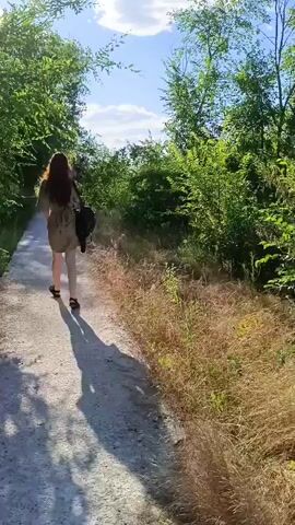 A person with long, dark hair walks away from the camera on a paved path surrounded by trees and vegetation. The person is wearing a light-colored dress and carrying a dark bag over their shoulder. The sun is shining, casting long shadows on the path. The sky is blue with some clouds.