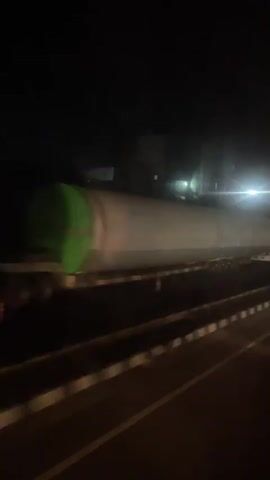 At night, a long, white cylindrical object, resembling an industrial tank, is being transported on a flatbed truck. The truck has a green section and is moving slowly on a road with visible road markings. Streetlights illuminate the scene, and the overall environment appears industrial. The video is taken from inside a car, showing the road ahead as the car follows the truck.