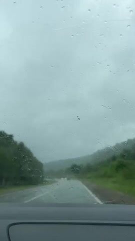 The video shows a view from inside a car driving on a wet road during a cloudy day. Raindrops are visible on the windshield. The road is surrounded by green hills and trees. There are other cars visible in the distance on the road. The windshield wipers are not visible in the frame. The weather appears to be overcast and rainy.