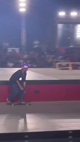 A skateboarder wearing a purple helmet and a dark shirt with blue jeans rides a skateboard on a ramp at night. The skateboarder is performing a trick near a set of stairs and a rail. There is a crowd of spectators in the background, and a person with a camera is filming the skateboarder. The event appears to be a skateboarding competition with banners and lighting.