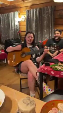 A woman with dark hair is sitting on a chair and playing a guitar. She is wearing a gray t-shirt, shorts, and colorful socks. A man with a beard sits at a table next to her, holding a guitar. They appear to be in a wood cabin with a table covered with a red patterned tablecloth. On the table, there's a plate with snacks and a bowl filled with sweets. The woman is singing, and the man is watching her. The cabin has log walls and curtains in the background.