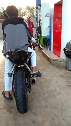 At a gas station, a person on a motorcycle waits for fuel. The motorcyclist is wearing a grey hoodie, white pants, and dark sandals. A gas station attendant, wearing a red uniform and a red cap, is seen in the background operating the fuel pump. The motorcycle has a black seat and a prominent exhaust pipe. The scene takes place on a paved surface, likely a gas station forecourt, with a fuel pump and a red building visible in the background. The lighting suggests it is daytime, possibly late afternoon due to the warm tones.