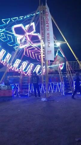 The video shows an amusement park ride at night. The ride is decorated with bright, colorful neon lights, including a large flower-like shape and geometric patterns. There is a tall central pole supporting the ride, with a banner attached to it that reads "PUTRA KAMPRET" and "PKR." Two sets of stairs lead up to the ride's platform, which is lined with more blue neon lights. A person wearing a dark jacket and pants stands on the ground in front of the ride, facing away from the camera. The ground appears to be dirt or gravel. The sky is dark, indicating it is nighttime. The ride is static in the video.