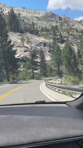 A first-person perspective video shows a car driving on a winding mountain road. The road has a double yellow line dividing lanes and a white line marking the edge. A metal guardrail is visible on the right side of the road. The surrounding environment consists of rocky, mountainous terrain with scattered pine trees. The sky is blue with white clouds. The sun appears to be shining. The dashboard of the car is visible in the foreground. No people are visible in the frame.