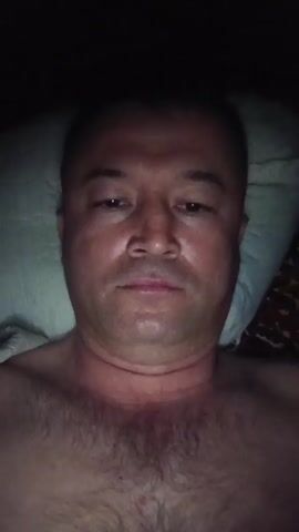A close-up, top-down shot shows a shirtless man lying on a pillow in a dark environment. The man has short, dark hair, and his face is the main focus. He initially makes a "peace" sign with his fingers before retracting them and looking straight ahead. He appears to be indoors, possibly in bed.