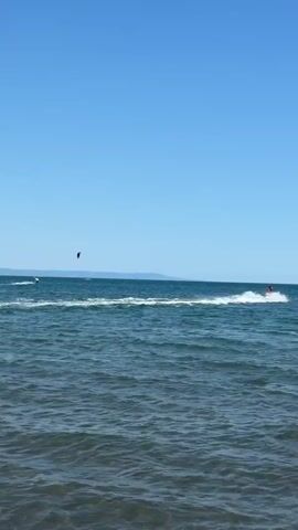 A static shot of a clear blue sky over a blue ocean. In the foreground, a person stands in shallow water, facing away from the camera. In the midground, there are several kite surfers and jet skiers. The kite surfers are in the process of riding the waves, some of them lifting their kites into the air. The jet skiers are creating wakes as they speed across the water. In the background, a distant coastline can be seen. The overall atmosphere is one of leisure and recreation on a sunny day.
