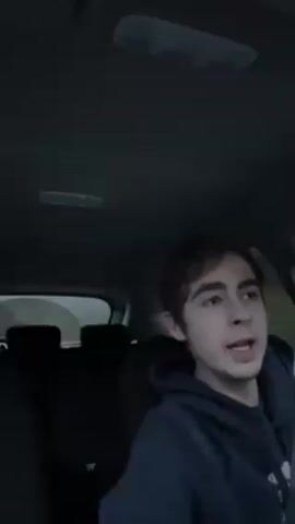 Here is a detailed description of the video:

A young man with brown hair is sitting in the driver's seat of a car. He is wearing a dark hoodie. The interior of the car is dark, and the view through the rear window is slightly blurred, suggesting the car is in motion. The man sings and then raises his clenched fist in the air as he speaks.