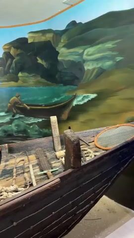 The video pans across a museum exhibit featuring a weathered wooden boat in the foreground, filled with nautical equipment like ropes, nets, and a barrel. Behind the boat is a large mural depicting a coastal scene with two figures in small boats and one on the rocky shore, engaged in fishing or handling nets. The mural shows a bright blue sky, a rocky coastline, and choppy water. The camera movement is slow and steady, highlighting the details of the boat and the painted backdrop.