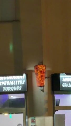 The video shows an exterior shot of a restaurant called ASYA, specializing in Turkish food. The sign reads "RESTAURANT SPECIALITES TURQUES". To the right of the entrance is another sign that reads "SANDWICH GRILL".  Above and between the two signs is a decorative light fixture shaped like a cylindrical column with an orange and red internal glow. The building is a light beige color. The scene appears to be at night.