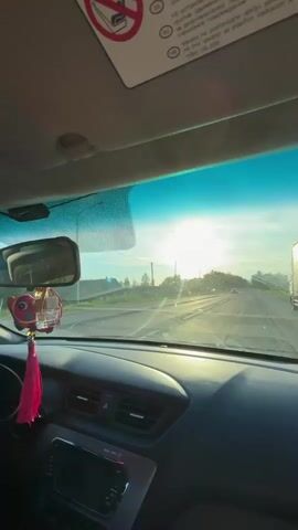 The video provides a first-person perspective from inside a car driving down a road. The bright sun is low in the sky directly ahead, creating a strong glare through the windshield and illuminating the dashboard. The road appears to be a two-lane highway or main road, with another car visible in the distance and a large white truck on the right side. Green trees and utility poles line the sides of the road.

Inside the car, a black dashboard is visible with air vents and a media control panel. A rearview mirror hangs centrally, and a pink, tasseled charm, possibly depicting a bird, dangles from it. Above the windshield, a sun visor is flipped up, displaying safety instructions in multiple languages, including Russian (RU) and Croatian (HR), warning against placing child seats on the passenger side due to airbags. The Russian text reads "НЕ УСТАНАВЛИВАЙТЕ детские удерживающие кресла на это сиденье" (DO NOT INSTALL child restraint systems on this seat), accompanied by an icon of a baby seat with a diagonal line through it. The sky is bright blue, hinting at either sunrise or sunset.