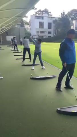 Several people are practicing their golf swings at a driving range. The driving range has a covered roof and is situated on a green, artificial turf surface. Each person stands on a black mat and hits golf balls into the distance. One man in a blue vest and dark pants is closest to the camera, holding a golf club. The others are further down the line, practicing their swings. In the background, there's a white building with large windows and trees. The weather appears to be sunny.