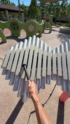 A person's hand is holding a black mallet and striking a large outdoor xylophone, which is made of many gray and white pipes. The xylophone is on a tan, rubberized surface, and in the background is a playground with green and yellow structures. The sun is shining, casting shadows.