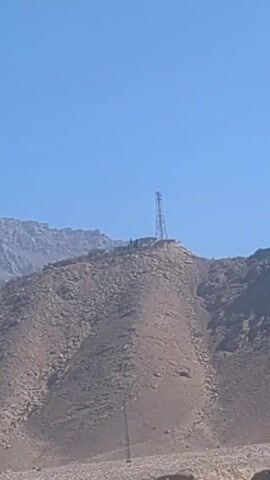 The video depicts a desert landscape under a clear blue sky. In the distance, there are mountains, one of which has a tall transmission tower on its peak. Nearer to the camera, there is a flat, barren area, and what appears to be a makeshift camp or settlement with tents or tarpaulins. The video is shot from a moving vehicle on a paved road, as seen by the road surface and concrete blocks along the side of the road.