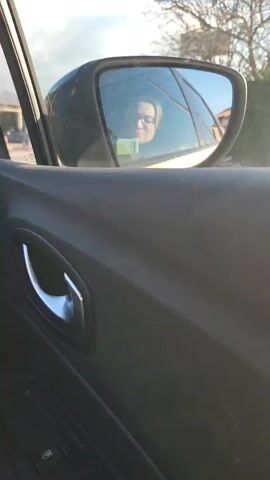 The video shows the interior of a car from the passenger side door. The door panel is visible, featuring a circular air vent, a silver door handle, and window controls. Outside the window, there are buildings, trees, and a billboard visible. The side mirror shows the reflection of a person with glasses, looking down at something in their hands. The camera seems to be stationary inside the moving car. The overall lighting is bright.
