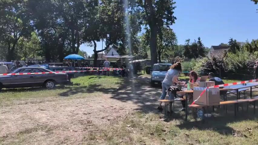 The video shows an outdoor gathering with several classic cars parked on a grassy area. A red and white ribbon cordons off sections of the area. In the foreground, there are picnic tables set up with people sitting at them. One person, wearing a grey t-shirt and jeans, is standing near a light blue car. Another car, a two-door sedan, is parked to the left of the frame. The environment is a sunny day with trees providing shade in the background. There are also some buildings visible in the distance. The overall atmosphere appears to be a casual event, possibly a car show or a community gathering.