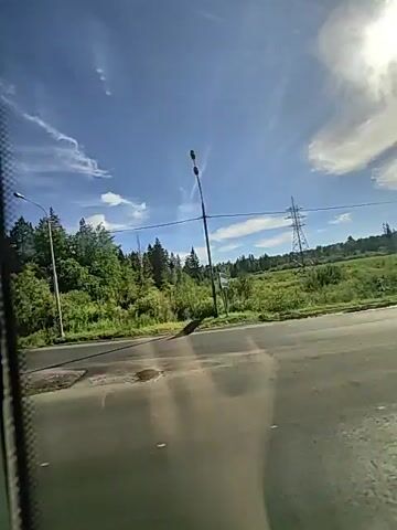 The video shows a view from a vehicle traveling on a paved road. On the left side of the road, there is a metal fence. Beyond the fence, there is a grassy area and then a dense line of trees. The sky is blue with some white clouds and bright sunlight. In the distance, there are power lines and a tall metal tower. The road appears to be a highway or a main road. There are streetlights visible along the side of the road. The time stamps indicate the video is very short, showing a continuous view of the passing scenery. No people are visible in the video.