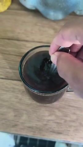 A close-up shot shows a hand stirring a dark liquid in a clear glass mug with a spoon. The mug is on a wooden surface. The liquid is swirling around the spoon.