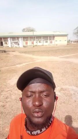 The video shows a man taking a selfie. The man is wearing a black baseball cap and an orange shirt with writing around the neck. He has a short beard and is wearing red earphones. Behind him are several low, single-story buildings with light-colored walls and dark roofs. The ground is dry and dirt. A man is sitting outside of one of the buildings. The sky is blue and the sun is shining.
