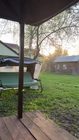 The camera pans across a serene backyard scene on a sunny day. In the foreground, a wooden porch is visible, with a swing set partially in view to the left.  Beyond the porch, a green lawn stretches out towards a wooden cabin and a small shack in the background. A ladder is leaning against the cabin wall. Several trees are scattered throughout the yard, some blossoming with white flowers. The sun is setting, casting a warm glow across the landscape. The sky is a gradient of soft blues and oranges.  A metal frame structure is seen in the right of the lawn.
