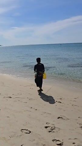 A video shows a sandy beach with footprints. Two individuals are present. One person, wearing a dark shirt and pants, stands on the sand near the water's edge, holding a fishing rod and a yellow bucket. Another person, in a red shirt and shorts, walks along the beach away from the camera. The ocean is calm with clear blue water, and the sky is blue with scattered clouds. The sun is shining, casting shadows on the sand.