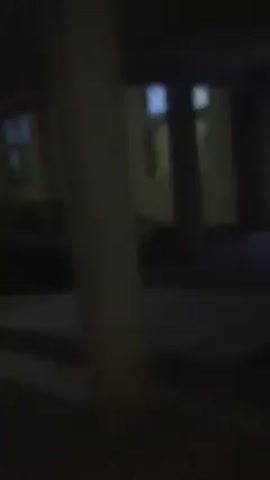 The video shows a first-person perspective moving through a dimly lit outdoor walkway with concrete pillars. The walkway leads to an open doorway that reveals a long, brightly lit indoor hallway with multiple doors on either side. The floor is light-colored, and the walls are a muted yellow. The light source in the hallway appears to be fluorescent. No people or other notable objects are visible. The camera movement is slightly shaky, suggesting it is handheld.