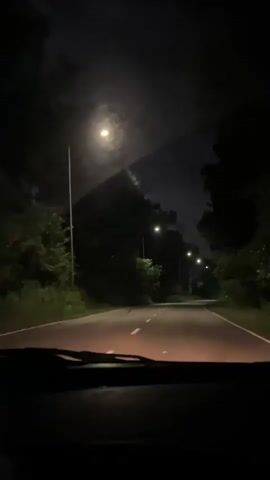 The video shows a first-person view from inside a car driving on a road at night. Streetlights illuminate the road, which is lined with trees on both sides. The sky is dark and appears cloudy. The headlights of the car are on, casting light on the road ahead. The video is dark overall, with the streetlights providing the main source of illumination.