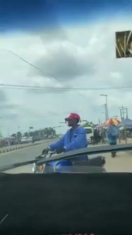 The scene is captured from inside a vehicle looking out through the windshield. A man on a blue and black motorcycle is stationary, positioned in the middle of the road. He is wearing a blue jacket, gloves, and a red cap. The background reveals a street scene with buildings, streetlights, and other people. There is a pedestrian walking along the side of the road to the right of the motorcycle. The sky above is blue with scattered clouds. The wipers of the car are visible in the foreground, along with the dashboard. The scene suggests a potentially busy street with normal daytime activity.