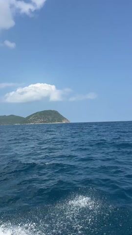 The video depicts a view from a boat on a calm, blue sea under a clear, blue sky dotted with scattered white clouds. A small, green, hilly island is visible in the distance to the left. The water is slightly choppy, with small waves across the surface. A metallic railing is visible at the bottom edge of the frame.