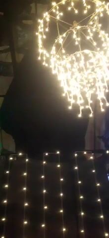 The video shows an outdoor scene at night, likely a religious festival or celebration. Numerous green and yellow lights are strung across trees and a railing. In the background, there is a large statue, possibly of a deity, decorated with garlands. A person is visible in the foreground, standing and facing the statue. The overall atmosphere appears festive and illuminated.