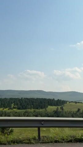 The video shows a view of a rural landscape from a moving vehicle. In the foreground, a metal guardrail runs horizontally across the frame. Beyond the guardrail, a green grassy slope with scattered trees leads to a dense forest. Further in the distance, rolling hills covered in a lighter green vegetation and sparse trees extend to the horizon. The sky is a clear blue with a few scattered white clouds. The camera appears to be positioned to the right of the roadway, looking towards the landscape. The time stamp indicates the video is approximately 5 seconds long.