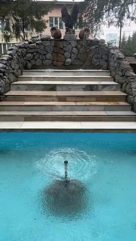 An outdoor water feature is captured, showcasing a decorative stone structure with multiple steps. At the pinnacle of the structure, a prominent statue of an eagle with outstretched wings is visible, flanked by two terracotta-like pots. Water cascades down the stone steps, creating a gentle waterfall effect. Below the stairs, a blue-tiled pool contains a fountain nozzle actively spraying water upwards, generating ripples on the water's surface. Trees and a building are discernible in the background. The scene appears to be set during the daytime under an overcast sky, lending a soft, diffused light to the environment. This tranquil setting is located at Sanatoriya Yumatovo Imeni 15-Letiya Bassr, Russia. No visible text is present.