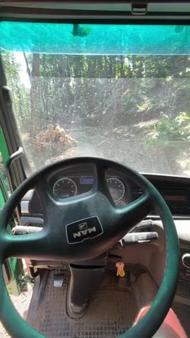 The video begins from the perspective of someone inside the cab of a green truck. The steering wheel, dashboard, and pedals are visible. Through the windshield, a dirt road leads uphill into a wooded area, with a pile of cut logs visible on the left. The camera then pans to the right, showing the truck door opening. The scene outside the truck is a forest with a steep, uneven terrain. The camera continues to pan, revealing more of the wooded environment and the path ahead. The video concludes with a view of the forest from the open truck door, showing trees and the unpaved road.